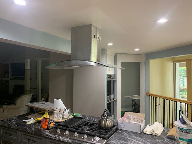 A kitchen island with a gas cooktop, overhead stainless steel vent hood, marble countertop, and some scattered items.