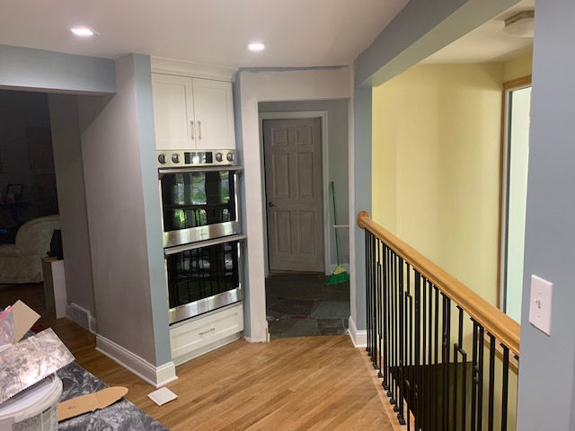 A stainless steel double wall oven in a white cabinet, set beside a staircase with wood railing and light-colored walls.