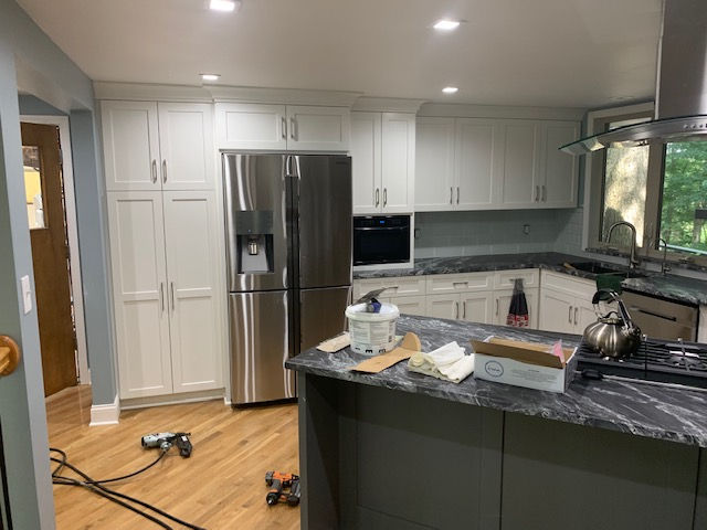 A modern kitchen featuring white cabinets, a stainless steel refrigerator, and a dark stone-topped island during renovation.