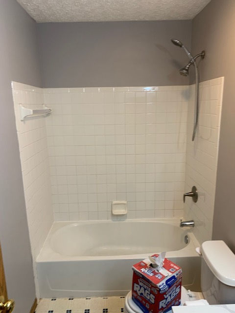 A bathroom featuring a white bathtub with a tiled surround, a chrome shower head, and a box of cleaning rags on the floor.