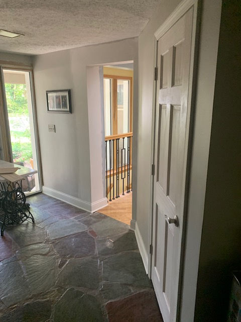 A slate-floored hallway with a light gray wall, a white six-panel door on the right, and a doorway showing a wood staircase.