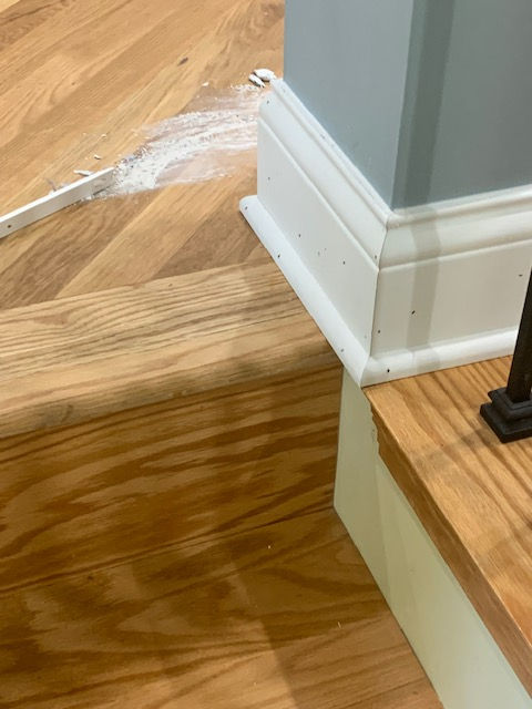 A white powdery substance spilled on a hardwood floor next to a white baseboard and the base of a staircase.