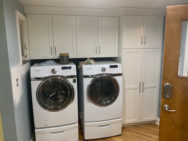 White washer and dryer with storage pedestals below, beneath white wall cabinets and next to a tall pantry cabinet.
