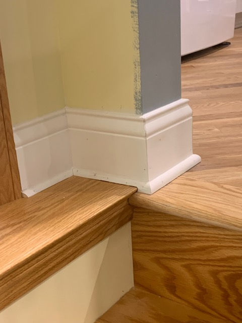 A white baseboard meets a wooden step corner, showing unfinished drywall tape on the adjacent wall corner.
