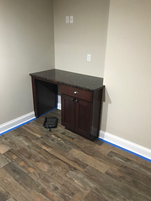 A built-in dark wood desk with a granite countertop sits in the corner of a room with light walls and wood-look flooring.
