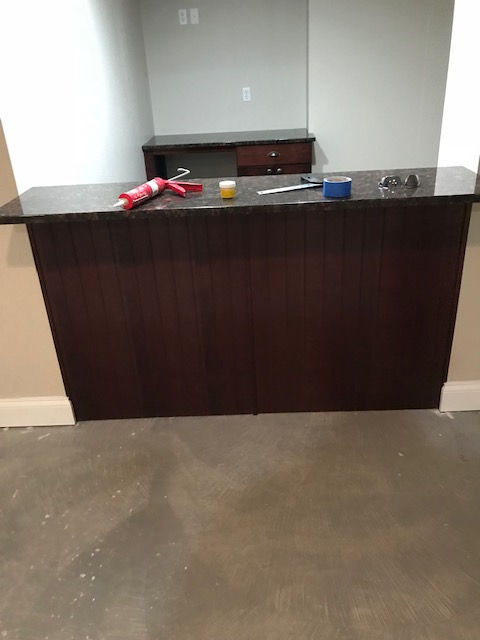 A dark wood bar counter with a granite top in an unfinished room, featuring a caulk gun, small jar, and tape roll on top.