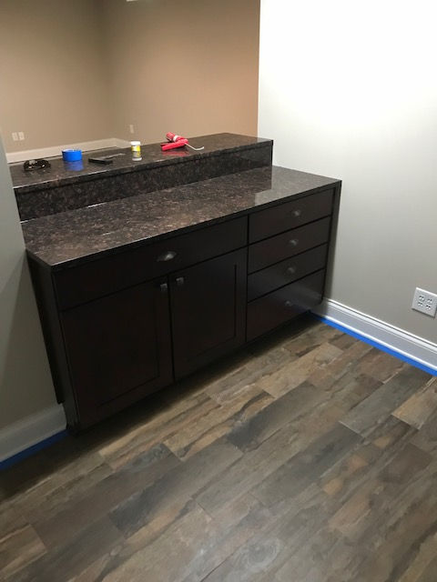 Dark brown kitchen cabinets with granite countertops installed against a beige wall, situated on light-toned wood flooring.