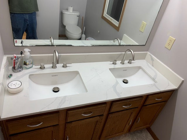 A double vanity bathroom sink with a white countertop and wooden cabinets, reflected in a large mirror.