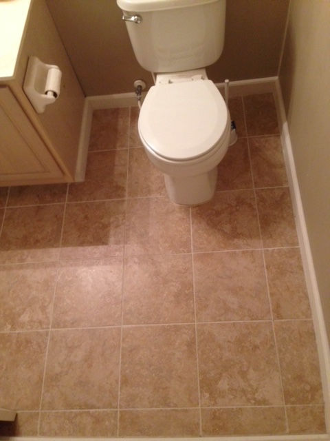 A top-down view of a small bathroom featuring a white toilet on a tan, square-tiled floor next to a beige vanity.