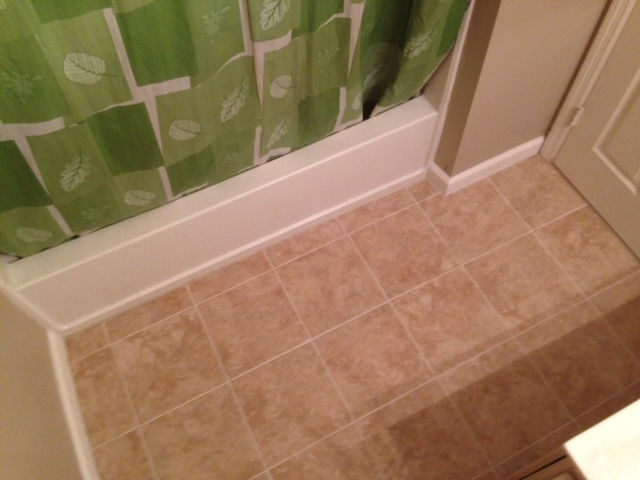 A high-angle shot of a beige tiled bathroom floor next to a white bathtub with a green leaf-patterned shower curtain.