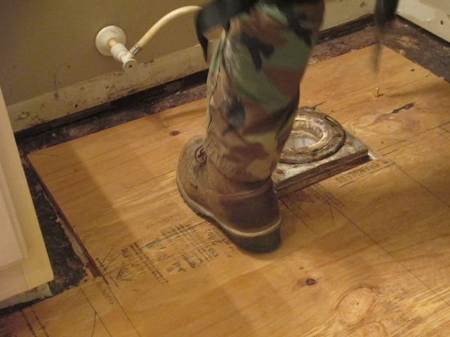 A boot stands on plywood flooring next to a toilet drain flange and a water supply line in a bathroom under renovation.