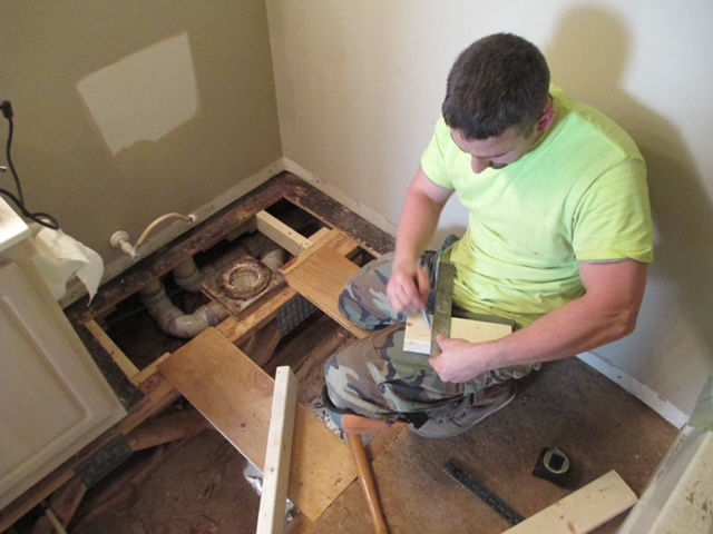 A person in a neon green shirt kneels on a subfloor, using a speed square to measure wood for a bathroom repair.