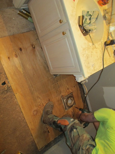 A person in work clothes uses a drill on a new plywood bathroom subfloor next to a vanity cabinet and toilet flange.