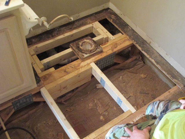 A bathroom floor undergoing renovation, showing exposed wooden joists around a toilet drain pipe and subfloor.