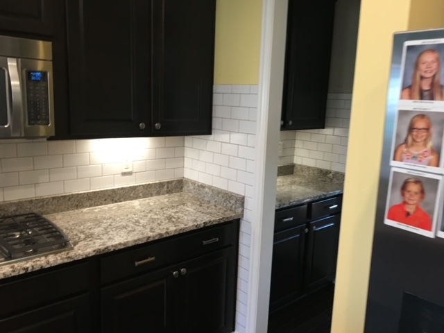 A kitchen with dark cabinets, granite countertops, and white subway tile backsplash, with framed photos on the side.