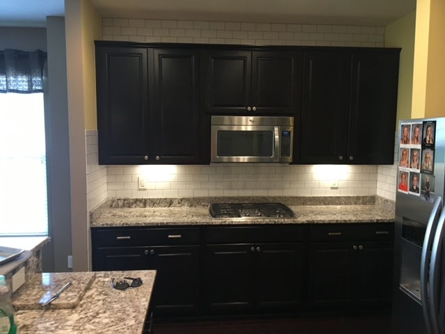 A kitchen with dark wood cabinets, granite countertops, a stainless steel microwave, and a white subway tile backsplash.