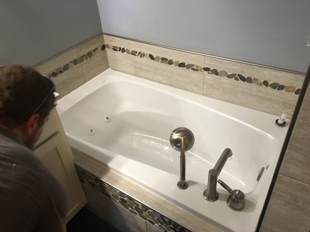 A person stands beside a modern white jetted bathtub featuring brushed nickel fixtures and decorative stone tile accents.