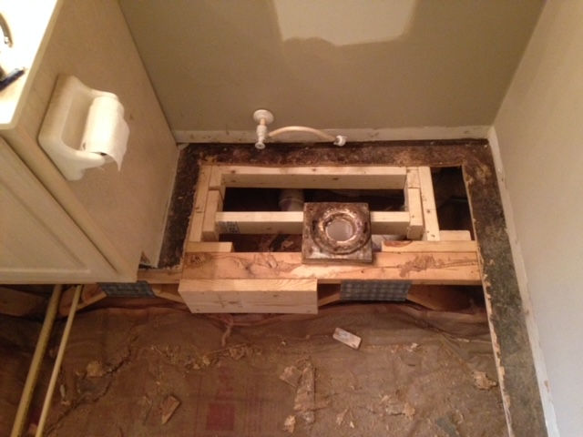 A bathroom floor renovation in progress, showing exposed subflooring, wooden joist framing, and a central toilet flange.