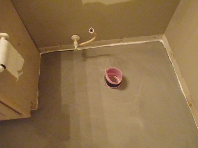 A small bathroom floor prepared for tiling with a toilet flange in the center and a water supply line emerging from the wall.