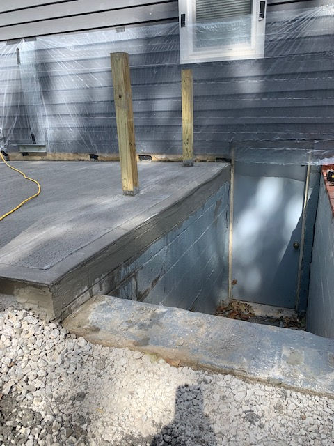A newly poured concrete patio level with a house foundation, featuring two wooden posts and an open basement stairwell.