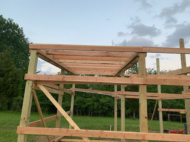 A wooden frame structure for a small building under construction, set in an open field against a cloudy sky.