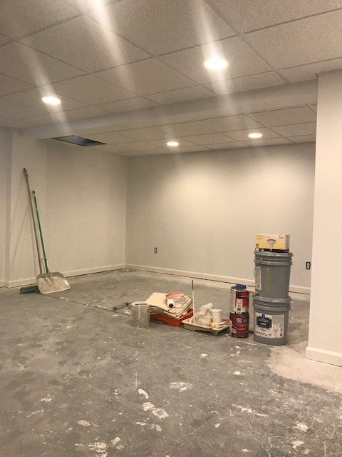 An unfinished basement room with white walls, a drop ceiling, a concrete floor, painting supplies, and a broom.