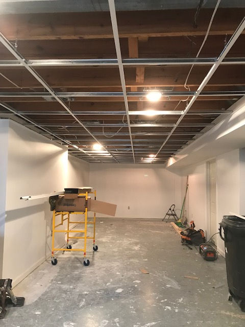 An unfinished basement room with white walls, concrete floors, a metal drop-ceiling frame, and construction equipment.