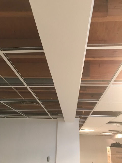 A partially installed drop ceiling with white metal grid segments and wooden beams visible beneath a white finished beam.