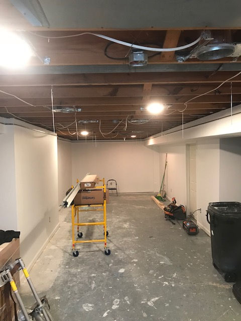 An unfinished basement room with exposed wooden ceiling joists, white painted walls, a yellow rolling scaffold, and tools.