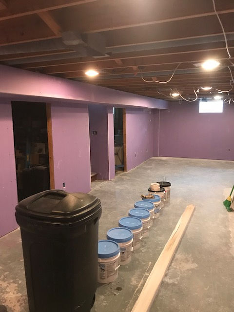 A partially finished basement with light purple walls, exposed wooden ceiling beams, and a row of paint buckets on the floor.