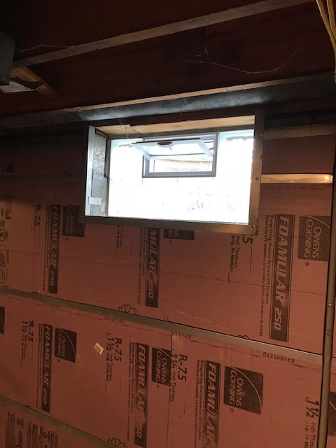 A small, hopper-style window set into a basement wall insulated with pink foam board panels.