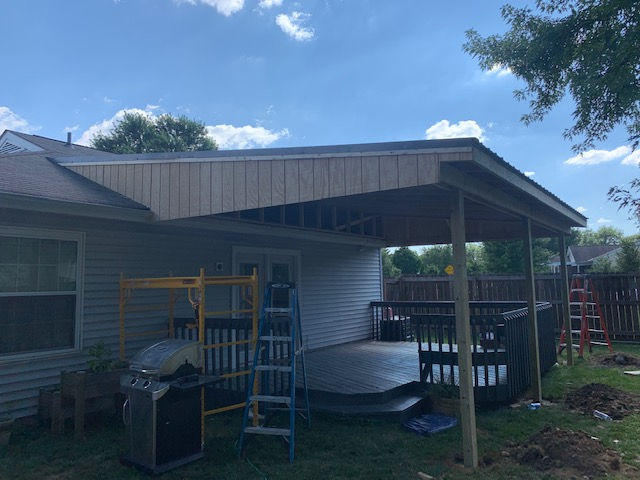 A partially finished wooden patio cover attached to a house with a deck, scaffolding, and ladders visible below.