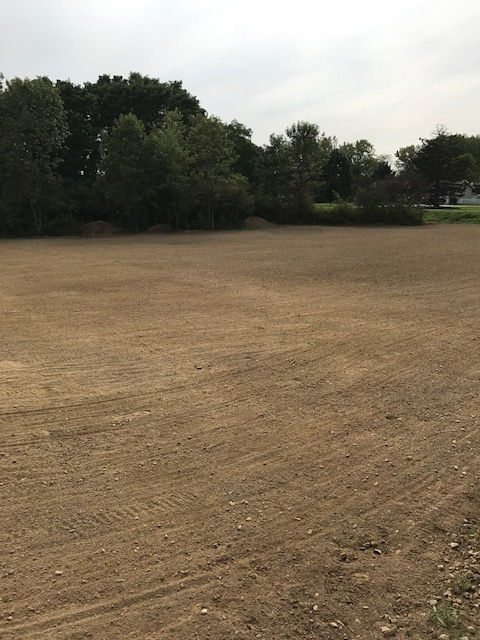 A large, empty field of tilled brown soil under a cloudy sky, bordered by a line of trees in the background.