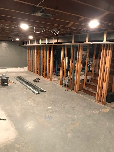 A basement under renovation with exposed wooden wall framing, unfinished ceiling, and a concrete floor.