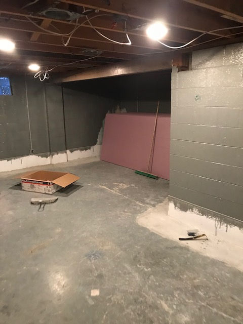 An unfinished basement with gray painted concrete block walls, exposed ceiling joists, and a sheet of pink insulation.