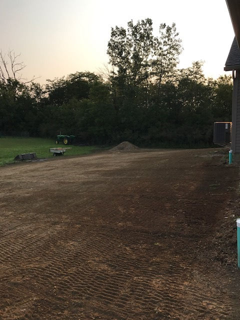 An open, bare dirt area or construction site during sunset, with a tree line and a small shed in the background.