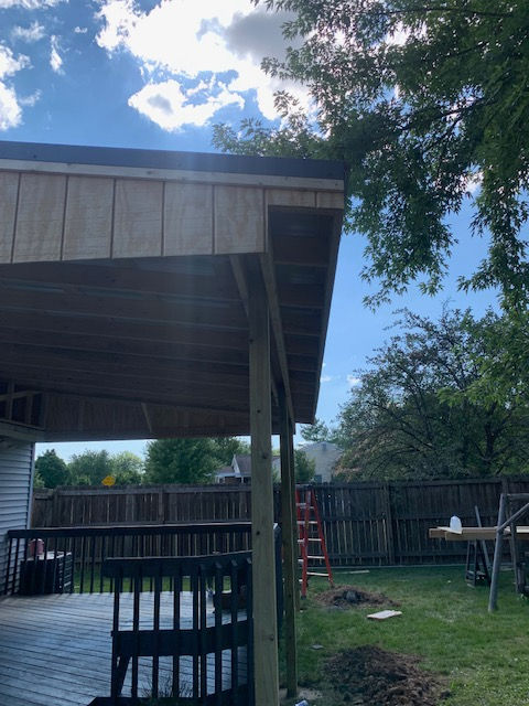A wooden porch roof structure under construction, viewed from a deck against a bright sky with trees and a fence nearby.