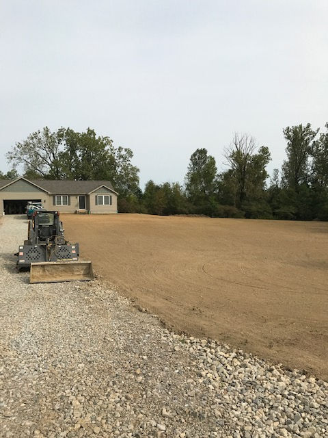 A skid steer parked on a gravel driveway next to a newly graded, bare dirt yard in front of a house.