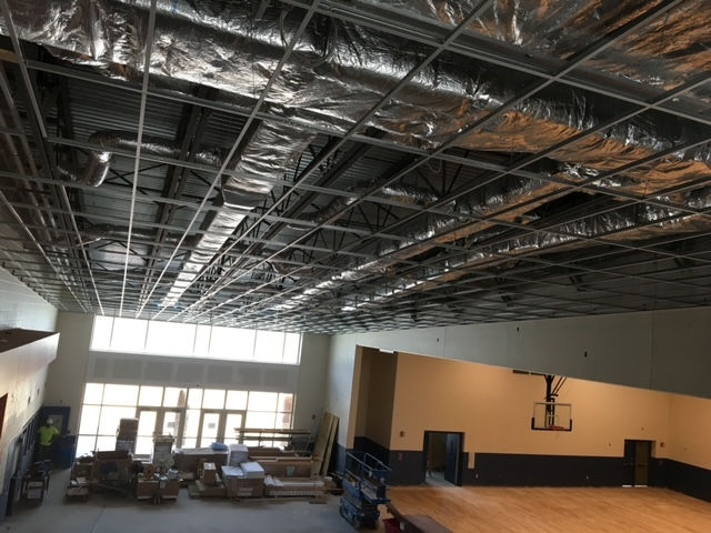 Interior view of a gymnasium under construction, showing an exposed grid ceiling, ductwork, and a basketball hoop.