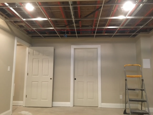 A basement room with two white doors, tan walls, a step ladder, and an exposed metal drop-ceiling grid above.