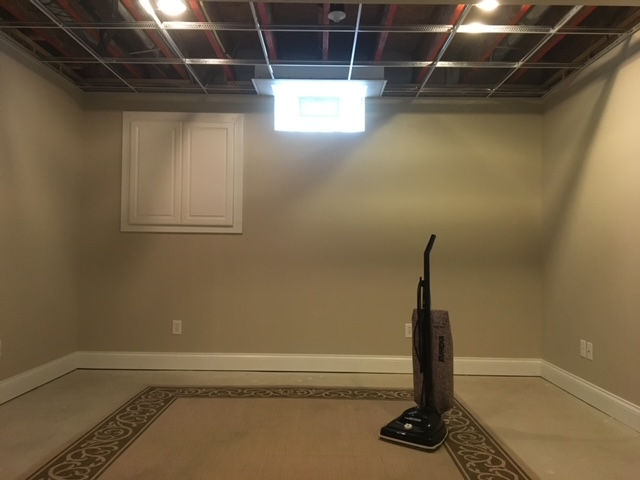 An empty basement room with tan walls, an unfinished ceiling grid, a window, a rug, and an upright vacuum cleaner.