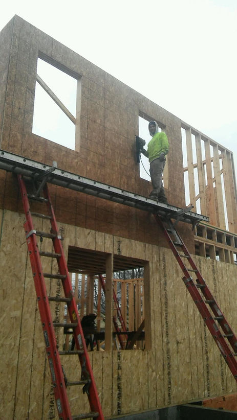 A construction worker in a high-visibility yellow vest stands on a metal platform to nail plywood onto a wood frame wall.