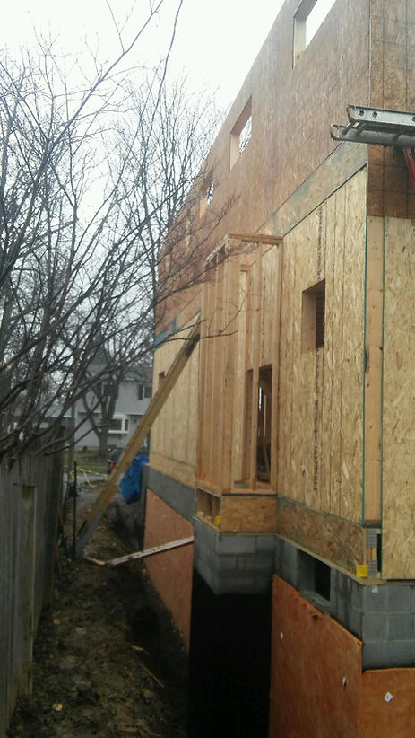 Side view of a house under construction with a cantilevered wall section supported by a wooden brace over a trench.
