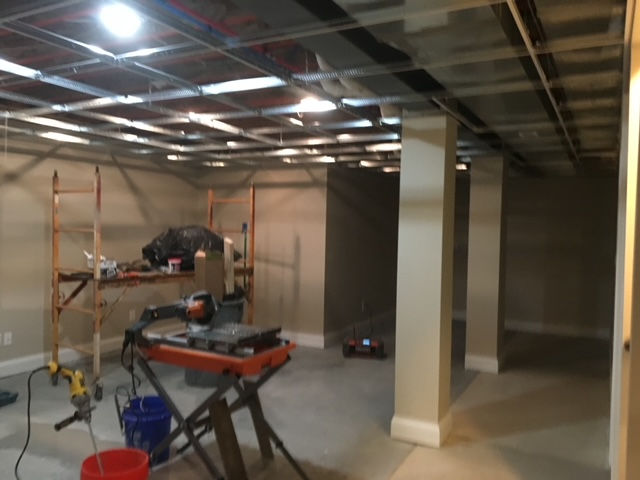 A basement under construction with a metal ceiling grid, support pillars, and various tools and equipment scattered around.