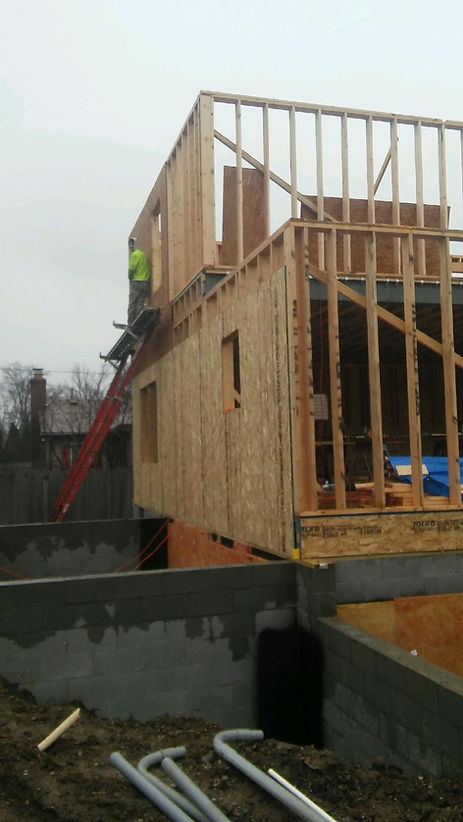 A wooden building frame under construction, featuring multiple levels, exposed studs, and a red ladder on a concrete base.