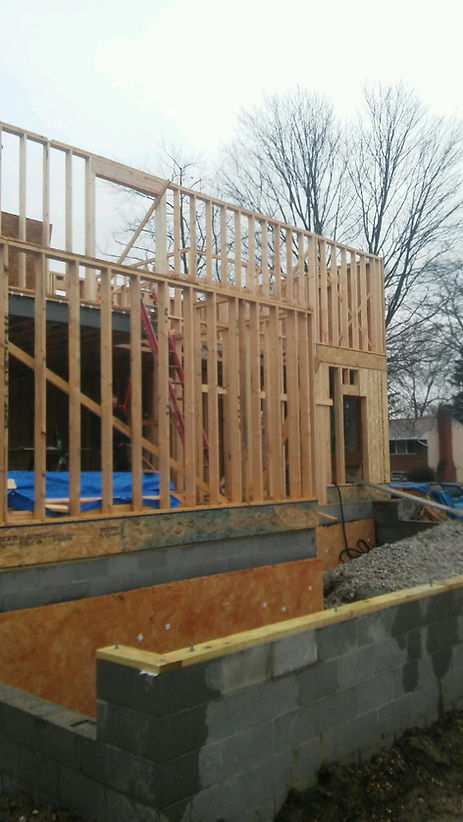 A construction site featuring an unfinished wooden house frame atop a concrete block foundation under an overcast sky.