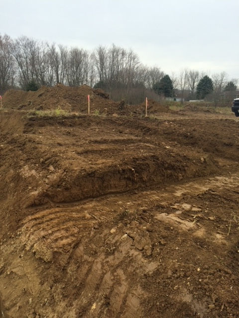 An excavated construction site showing layered brown soil, dirt mounds, and wooden stakes against a background of trees.