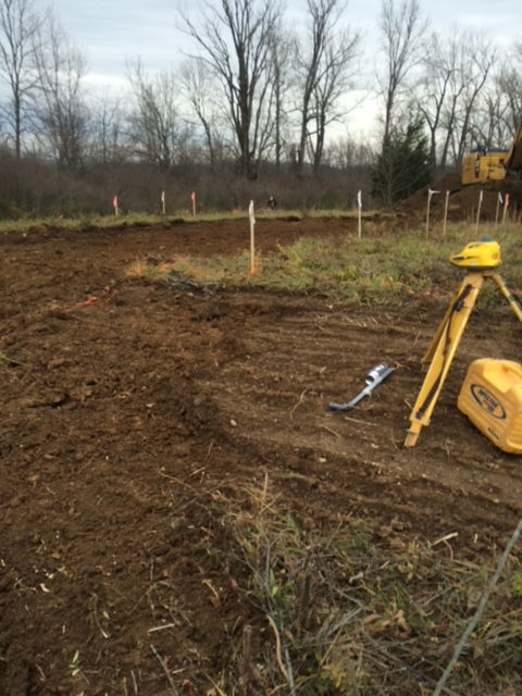 A yellow laser level on a tripod stands in a cleared dirt field with wooden stakes marking a property line.
