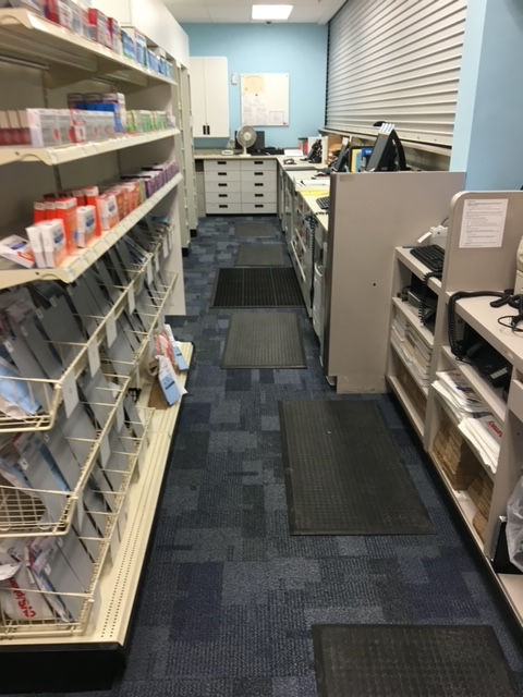 An interior view of a narrow pharmacy workspace with shelving units on the left, a counter on the right, and floor mats.