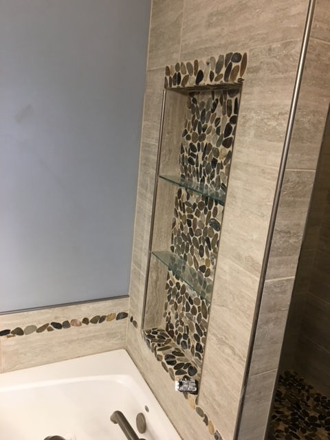A bathroom niche with glass shelves and decorative pebble tile inlay, set into a beige tiled wall above a white bathtub.